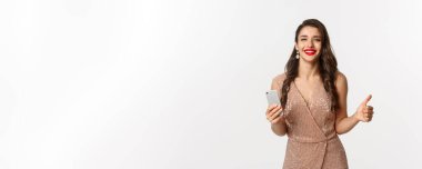 Tatiller, online alışveriş konsepti. Zarif elbise ve mücevher giymiş çekici bir kadın. Onaylı baş parmağını gösterirken cep telefonu kullanıyor. Gülümsüyor..