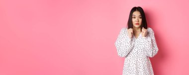 Korkmuş Asyalı kız endişeli görünüyor, korkmuş ve gergin bir şekilde kameraya bakıyor pembe arka planda duruyor..