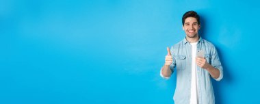 Çevrimiçi alışveriş, uygulama ve teknoloji kavramı. Normal giyinmiş, gülümseyen, akıllı telefon uygulaması kullandıktan sonra onay veren, mavi arka planda duran tatmin olmuş bir adam..