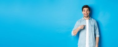 Mutlu ve şaşkın adam kendini işaret ediyor, gülümsüyor, mutlu, mavi arka planda duruyor..