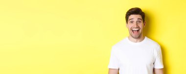 Harika haberlere tepki veren, beyaz tişörtlü, sarı arka planda duran, şaşırmış yakışıklının yakın çekimi..