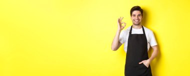 Siyah önlüklü, gülümseyen genç barista, tamam işareti gösteriyor, kahve ya da restoran öneriyor, sarı arka planda duruyor..