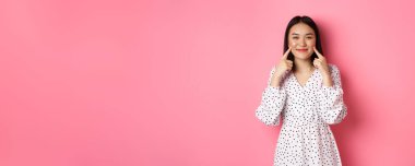 Tatlı, utangaç Asyalı kız yanaklarını dürtüyor, kameraya mutlu bir şekilde gülümsüyor, romantik beyaz elbiseler giyiyor, pembe arka planda duruyor..