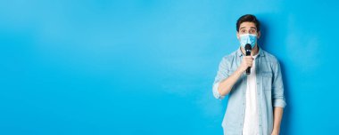 Tıbbi maskeli genç adam konuşma yapıyor, mikrofonu tutuyor ve kafası karışmış görünüyor, mavi arka planda duruyor..