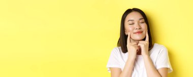 Güzellik ve cilt bakımı. Kısa siyah saçlı, sağlıklı, parlak tenli, gülümseyen ve yanaklarındaki gamzelere dokunan, sarı arka planda duran Asyalı genç bir kadın..