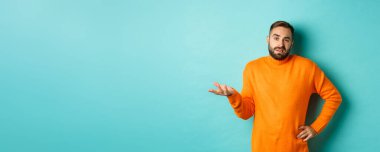 Dikkatsiz ve umursamaz adam omuz silkiyor, rahatsız edilmemiş görünüyor, hiçbir şey bilmiyor, açık mavi arka planda duruyor..