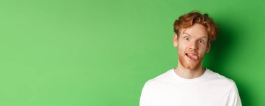 Duygular ve moda konsepti. Komik kızıl saçlı adamın aptal suratlar sergilemesi, dili yapışması ve kameraya bakması, yeşil arka planda durması..