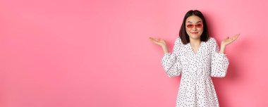 Kameraya gülümseyen sevimli Asyalı kadın turist, omuz silkiyor, habersiz, modaya uygun güneş gözlüğü takıyor ve beyaz elbise giyiyor, pembe arka planda duruyor..