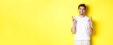 Umut dolu gülümseyen adam parmaklarını çapraz yapıyor, dilek diliyor ya da dua ediyor, sarı arka planda duruyor..