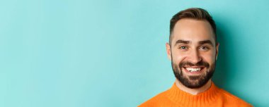 Kameraya gülümseyen, turkuaz arka planda turuncu süveter giyen, sakallı, yakışıklı beyaz adamın vesikalık resmi..