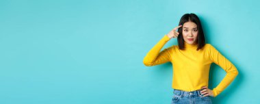 Are you stupid. Portrait of skeptical asian woman scolding someone crazy or dumb, pointing finger at head and staring at camera with dismay, blue background.