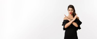 Moda ve güzellik. Siyah elbiseli ciddi ve kendinden emin bir kadın model, çapraz işaret ve somurtma gösteriyor, jesti durdur, hayır diyor, beyaz arka planda duruyor..