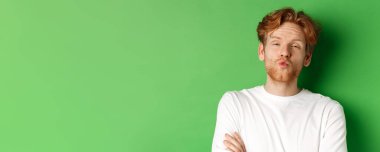 Duygular ve moda konsepti. Aptal kızıl saçlı, sakallı, dudaklarını büzüştüren ve öpüşmek için eğilen yeşil arka planda duran adam..