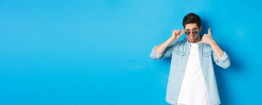 Güneş gözlüklü yakışıklı maço adam seninle flört ediyor, telefon işareti yapıyor ve göz kırpıyor, bana jest diyor, mavi arka planda duruyor..