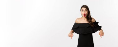 Moda ve güzellik. Siyah elbiseli zarif bir kadın, parmakları aşağıda, tanıtım yapıyor ve gülümsüyor, beyaz arka planda duruyor..