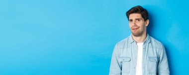 Kafası karışık ve rahatsız bir adam garip ya da ürpertici bir şeye bakıyor, kötü reklamdan korkup, mavi arka planda duruyor..