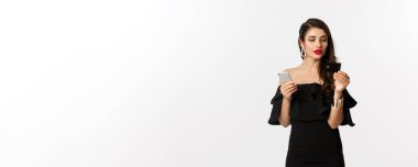 Moda ve alışveriş konsepti. Makyajlı ve siyah elbiseli çekici bir kadın online, kredi kartı ve cep telefonuyla ödeme yapıyor, beyaz arka plan.