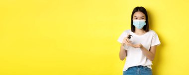 Covid, sağlık ve salgın hastalık konsepti. El dezenfekte etmek için el dezenfektanı kullanan, sarı arka planda duran, tıbbi maskeli tatlı Asyalı kadın..