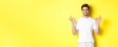 Rahatlamış adam gülümsüyor, tamam işaretleri gösteriyor, onaylıyor ya da kabul ediyor, sarı arka planda duruyor..