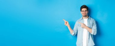 Gözlüklü hoşnutsuz ve şüpheci bir erkek model, kötü bir şeyi işaret ediyor, korkunç bir reklam veriyor, mavi arka planda duruyor..