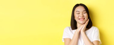 Romantik Asyalı kadının sevimli bir şeyin hayalini kurduğu yakın plan, gözlerini kapat ve yüzüne yakın el ele tut, güzel bir şey hayal et, sarı arka planda dur..