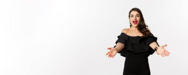Moda ve güzellik konsepti. Cazibeli siyah elbiseli mutlu genç bir kadın, elini uzatıp bir şeyler alıyor, sarılmayı bekliyor, beyaz bir arka plan..