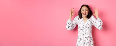 Hoş, genç Asyalı bir kadın iyi bir işaret veriyor, övgüler yağdırıyor ve onaylıyor, tatmin olmuş görünüyor, pembe arka plana karşı duruyor..