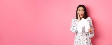 Gayrimenkul. Şaşırmış ve şaşırmış Asyalı kadın kameraya bakıyor, vay canına diyor ve kağıt ev modelini gösteriyor, pembe arka planda duruyor..