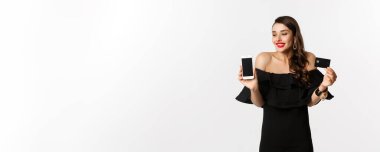 Moda ve online alışveriş konsepti. Siyah elbiseli mutlu genç bir kadın, kredi kartı ve mobil ekranı gösteriyor, beyaz arka planda duruyor..