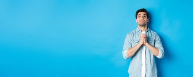 Umutlu genç adam yukarı bakıyor ve Tanrı 'ya dua ediyor, yardım için yalvarıyor, mavi arka plana karşı duruyor..