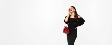 Güzellik ve moda konsepti. Siyah elbise, topuklu ayakkabı ve çantayla alışverişe çıkan, kendine güvenen, beyaz arka planda duran zarif genç bir kadın..