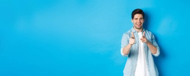 Küstah yakışıklı adam seni işaret ediyor, flört ediyor, mavi arka planda sıradan kıyafetler içinde dikiliyor..