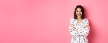 Bahar elbisesi giymiş güzel bir Asyalı kadın gülümsüyor, kolları göğsünde ve kendine güvenen pembe arka planda duruyor..