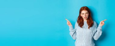 Süveter giyen kafası karışık bir kızıl kız parmaklarını yana doğru gösteriyor, kameraya şüpheyle bakıyor, mavi arka planda duruyor. Boşluğu kopyala