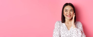 Utangaç Asyalı kadın, yanağına dokunuyor ve soluna bakıyor, pembe arka planda dikiliyor..