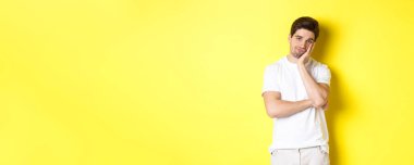 Sıkılmış ve keyifsiz bir adam, kayıtsız bakıyor, sarı arka planda beyaz elbiseler içinde duruyor..