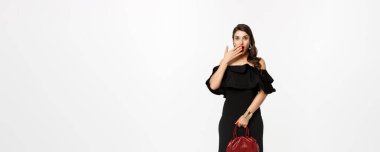 Güzellik ve moda konsepti. Siyah elbiseli, topuklu ayakkabı çantalı, kameraya bakan, şaşırmış, ağzı açık, beyaz arka planı olan bir kadın..