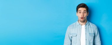 Yakışıklı, şaşırmış adamın yakın çekimi, ağzını aç ve hayret verici bir şekilde, anonsu duy, mavi arka planda dur..