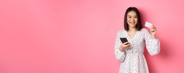 Çekici genç Asyalı kadın online sipariş veriyor, kredi kartı ve cep telefonu tutuyor, internet alışverişi yapıyor, pembe arka planda mutlu duruyor..