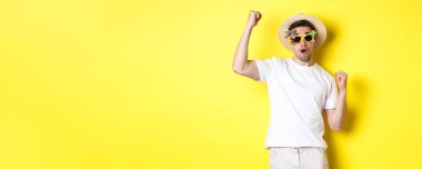 Turizm ve yaşam tarzı kavramı. Gezinin tadını çıkaran mutlu turist, seni destekliyor, yumruk tokuşturuyor ve başarıyor, yaz şapkası ve güneş gözlükleriyle yolculuğa çıkıyor, sarı arka planda..