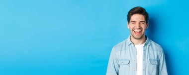 Mutlu, genç, sakallı, sıradan kıyafetler giymiş, gülen, neşeli, mavi arka planda duran bir adamın yakın çekimi..