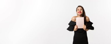 Moda ve alışveriş konsepti. Kırmızı dudaklı mutlu genç kadın, siyah elbise giyiyor, seviniyor ve dijital tablet tutuyor, ödül kazanıyor, beyaz arka plan.