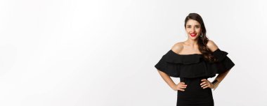 Kırmızı rujlu, siyah elbiseli, gülümseyen, beyaz arka planda duran güzel bir kadının belden yukarı pozu..