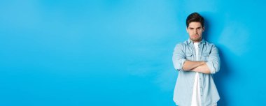 Öfkeli sakallı adam göğsünde kollarını kavuşturuyor, kaşlarını çatıyor, kızgın ve gücenmiş görünüyor, mavi arka planda duruyor..