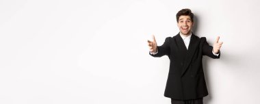 Takım elbiseli mutlu çekici bir adamın portresi, bir partiye ev sahipliği yapıyor, seni selamlamak için ellerini ileri uzatıyor, bir şey tutmak ya da sarılmak istiyor, beyaz arka planda dikiliyor..