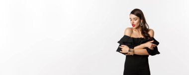 Güzellik ve moda konsepti. Siyah elbiseli, kırmızı dudaklı, şehvetli ve çekici bir kadın, kendini kucaklıyor ve nazikçe aşağıya bakıyor, beyaz arka planda duruyor..