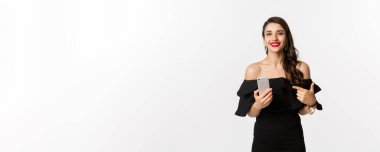 İnternetten alışveriş konsepti. Siyah elbiseli şık bir kadın, makyaj yapıyor, cep telefonunu işaret ediyor, gülümsüyor, beyaz arka planda duruyor..