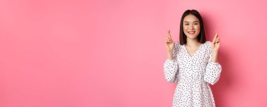 Tatlı Asyalı bir kadın dilek diliyor. Mutlu bir şekilde gülümsüyor ve şans diliyor. Pembe arka planda bekliyor..