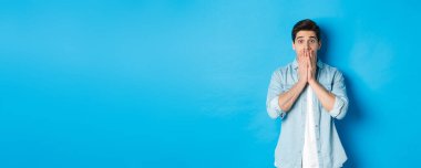 Şok olmuş yetişkin bir adamın görüntüsü kameraya hayranlıkla bakıyor, nefes nefese ve ağızlarını ellerle kapatıyor, mavi arka planda dikiliyor..