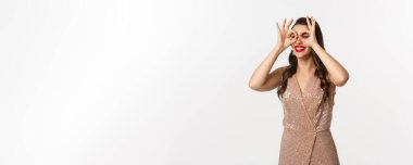 Noel partisi ve kutlama konsepti. Genç, güzel bir kadın mutlu, uzaktan bir şey görüyor, el dürbününden bakıyor, zarif bir elbise giyiyor, beyaz bir arka plan..